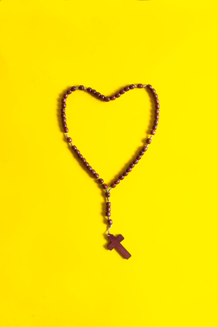 A heart-shaped rosary with a wooden cross placed on a vibrant yellow background.
